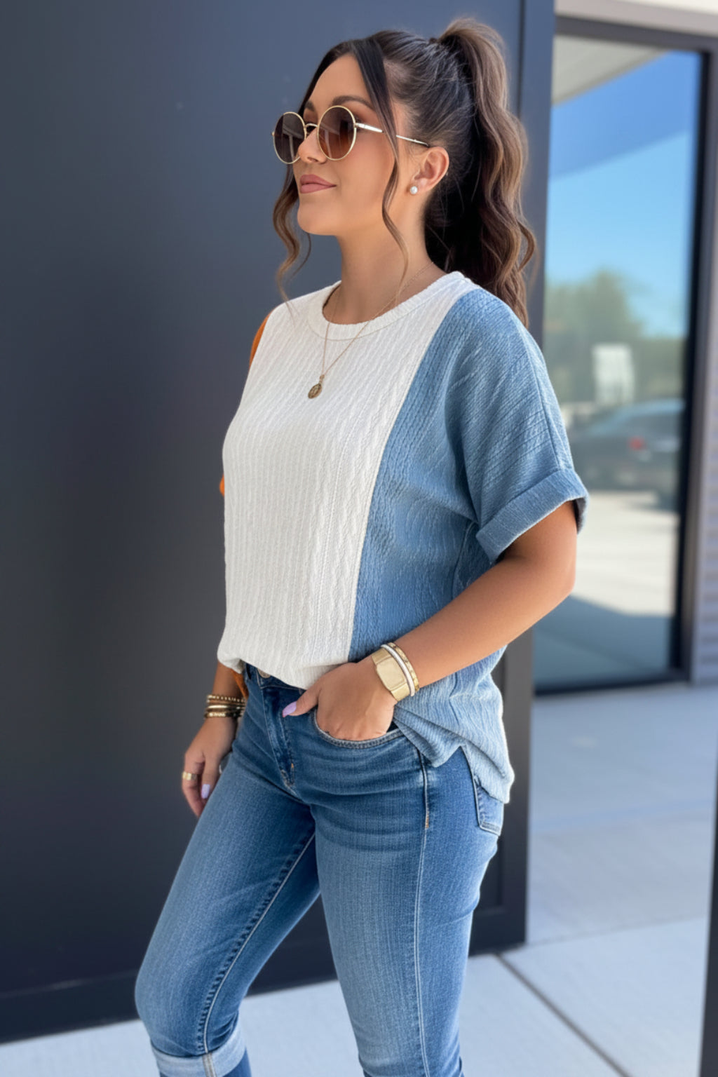 Woman wearing a white blouse with orange sleeves and denim shorts, standing against a blurred outdoor background.