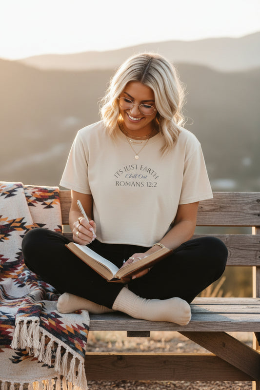A light beige short-sleeve crop top t-shirt with a graphic print on the chest reading 'IT'S JUST EARTH!' and 'ROMANS 12:2' in dark gray text, displayed flat against a white background.