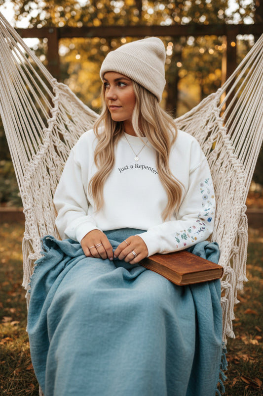 An off-white crewneck sweatshirt featuring the text 'Just a Repenter' printed in black on the chest and a vertical multicolor floral pattern down the right sleeve outside.