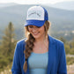 A blue and white trucker hat with a white front panel, light blue vertical stripes, and the words 'Homestead & Heart' in black script on the front. The brim and mesh back panels are royal blue.