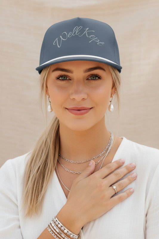 A slate blue snapback cap with 'Well Kept' embroidered in white script across the front, featuring a white stripe along the brim and a matching blue button on top.
